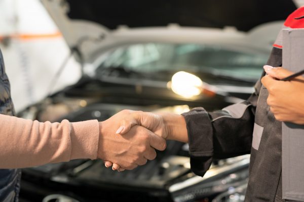 Young professional technician of car service shaking hand of client in workshop